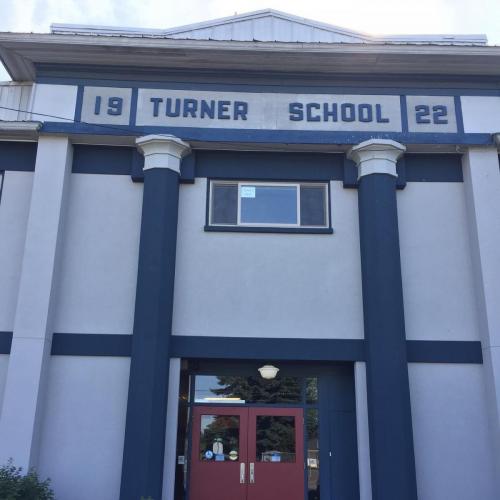 6. Location: Turner Elementary School. Image: A white, two-storey building with dark blue pillars on either side of the door, and dark blue trim where the floor and ceiling of the upper storey would be. The sign at the top says 19 Turner School 22.